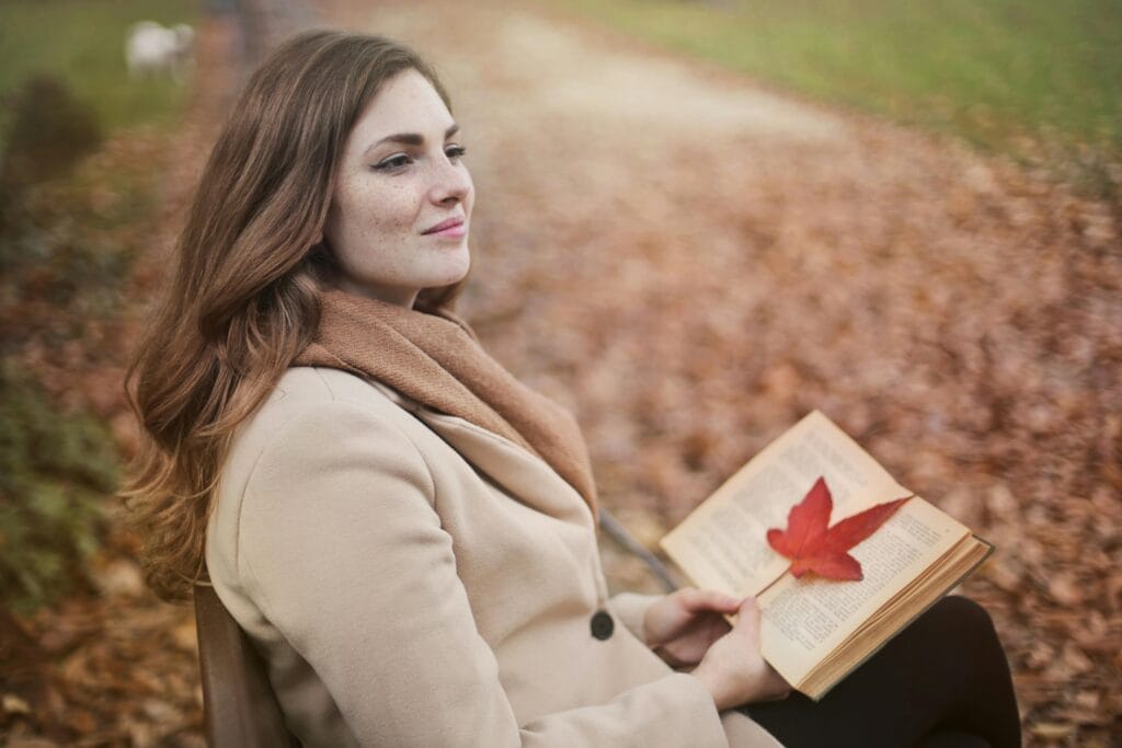 Vista lateral de una joven pacífica con un abrigo cálido y una bufanda sentada en un banco con un libro abierto en las manos y mirando hacia otro lado pensativamente mientras descansa en el parque de otoño