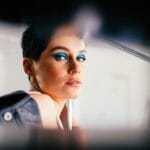 Vista lateral de una joven de autoestima con maquillaje brillante y cabello corto en una chaqueta de cuero de moda mirando a la cámara a través del agujero
