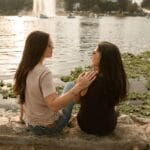 Dos mujeres se sientan junto a un lago, disfrutando de un momento de amistad y tranquilidad al aire libre.