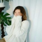 Una mujer con un suéter de punto blanco se cubre juguetonamente la cara con las manos en el interior.
