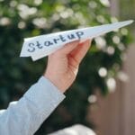 Primer plano de una mano sosteniendo un avión de papel con "Startup" escrito, simbolizando el lanzamiento de un negocio.