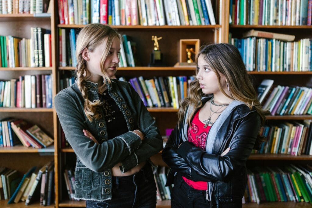 Dos adolescentes en una biblioteca con los brazos cruzados, de pie frente a estanterías llenas de libros.