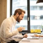 Hombre en la oficina con café, sonriendo mientras trabaja en la computadora portátil, captura la esencia del trabajo remoto.