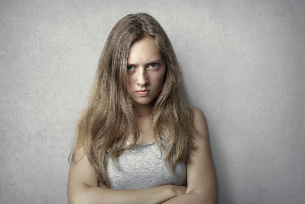 Retrato de una mujer feroz con los brazos cruzados y una expresión intensa sobre un fondo gris.