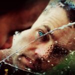 Un hombre de ojos verdes se asoma a un espejo agrietado y mojado, transmitiendo soledad y pensamiento.